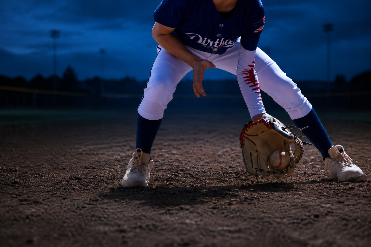 A close-up of a youth baseball player fielding a ground ball during blue-hour practice